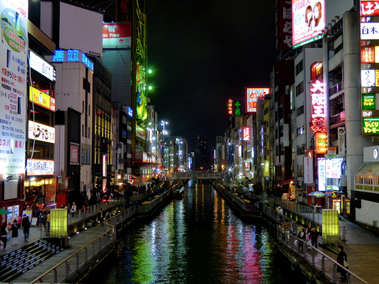A view looking out over a canal lined with multi-story buildings to both sides, their illuminated advertisements reflecting on the water