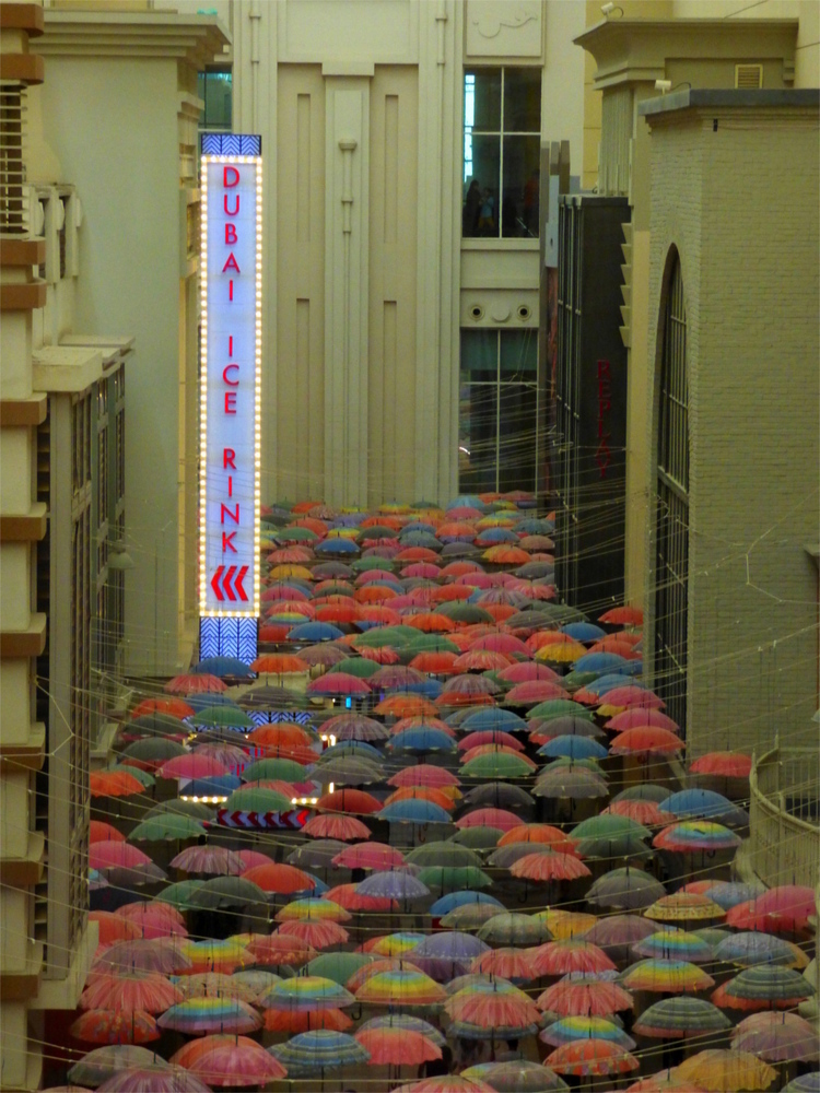 Interior of a sandstone-coloured mall with colourful umbrellas suspended from the ceiling to provide an outside-atmosphere, photographed from above. A vertical sign reads 'Dubai Ice Rink'.