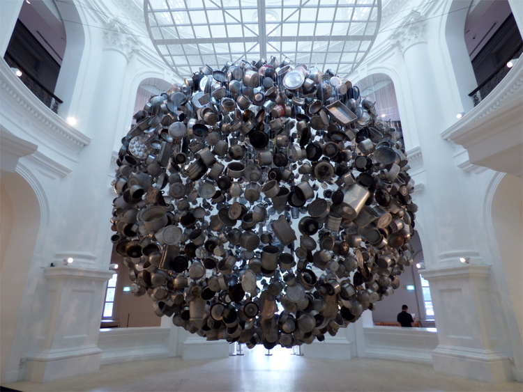 An art installation in a bright white foyer consisting of metal cooking utensils suspended from the ceiling in the form of a perfect sphere