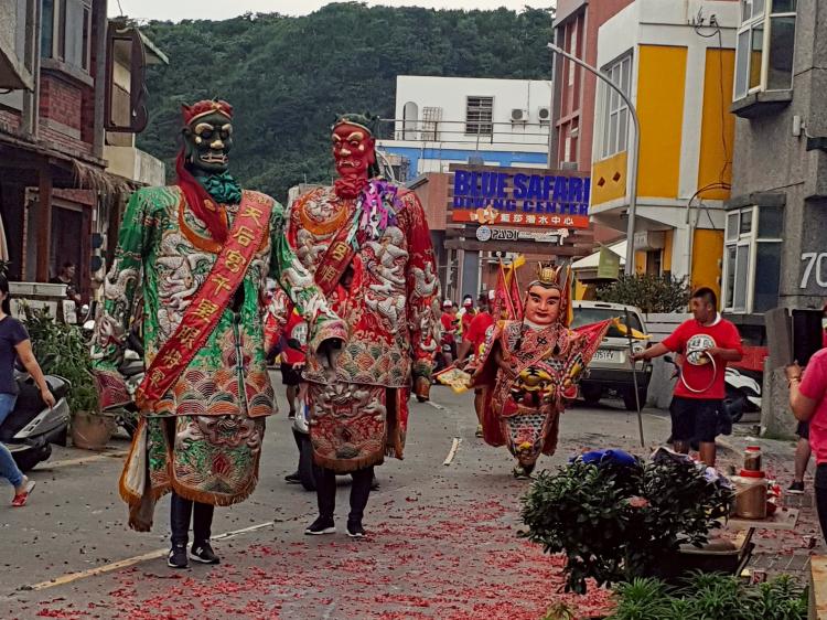 Two street performers wearing colourful, larger-than-life costumes walking on a street littered with burnt-out fireworks