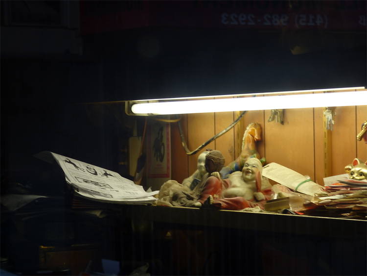 A small porcelain figure showing a resting Buddha laying under a fluorescent tube surrounded by stuff in an otherwise dark room