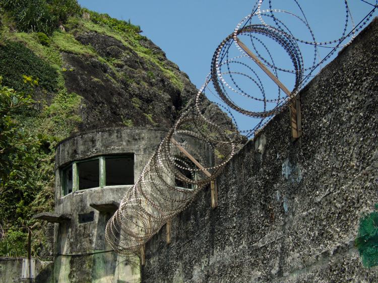 Barbed wire running along the top of a concrete wall leading to a round, abandoned-looking watchtower