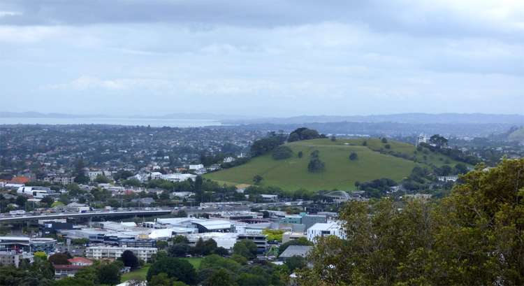 A cone-shaped green hill with the hint of a crater on top emerging from a suburban city landscape