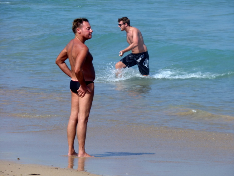 A slightly sun-burnt older man in speedos standing on the shore looking out at the sea contently