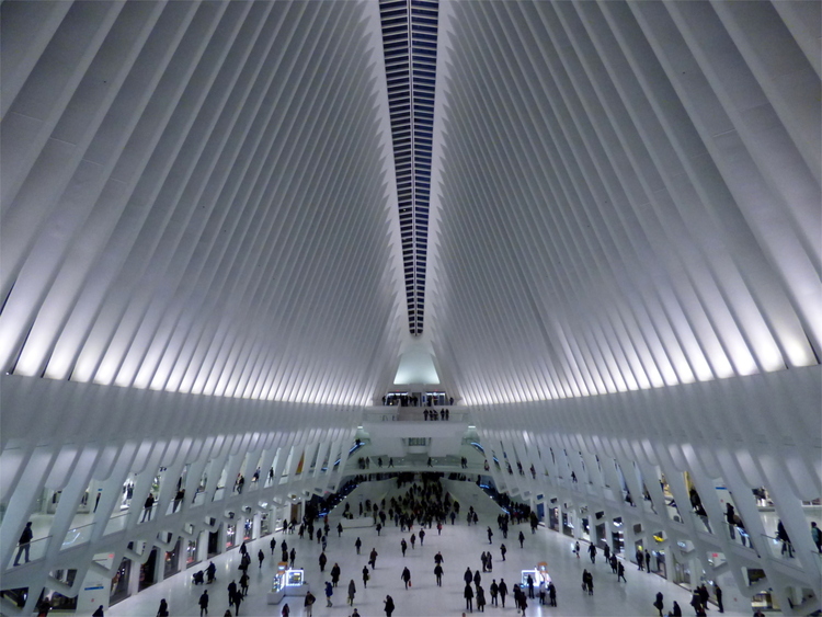 The tall, plain white interior space of a mall with a ceiling construction reminiscent of an alien ribcage and people walking across the floor like ants