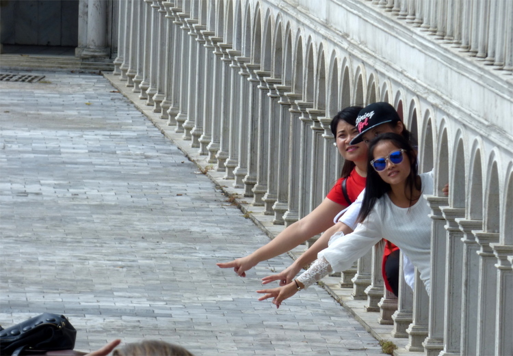 Three people poking out from arches in the miniature replica of an Italian square, making peace-signs at the camera