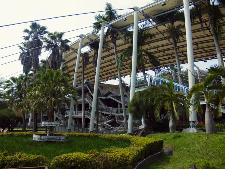 Remains of a collapsed building preserved under a metal roof construction surrounded by a landscaped park