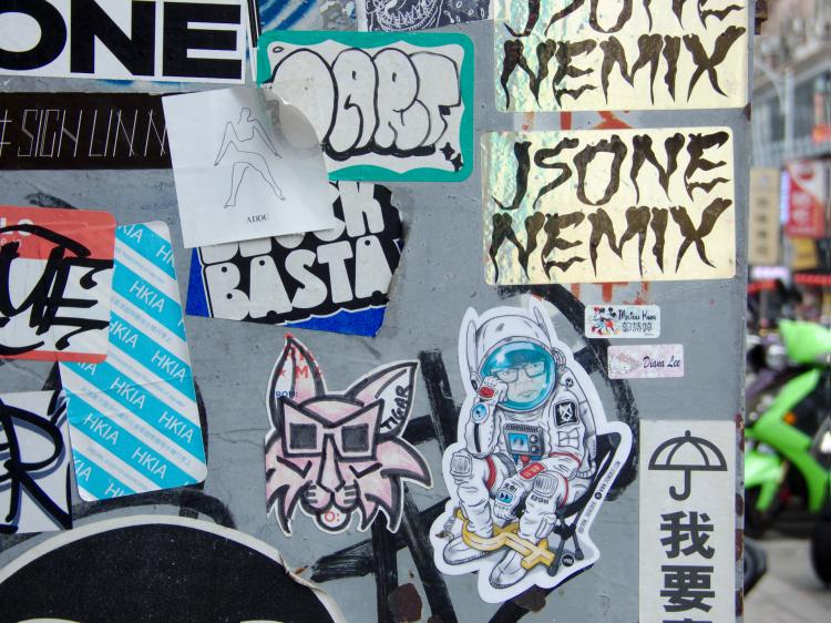 Stickers on the back of a street sign, one showing a contemplative astronaut, another a cat with 3D-glasses