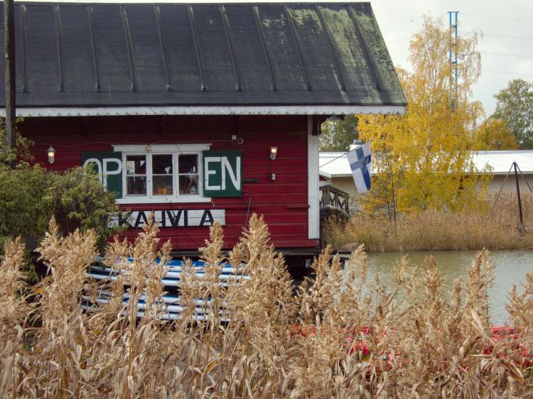 A small red wooden cabin on a lake, viewed through some tall brown reeds