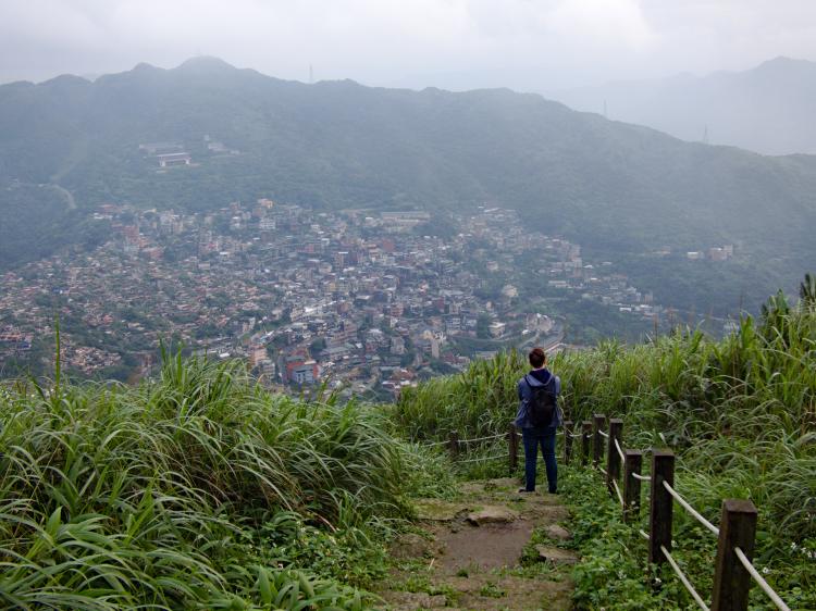 Jan standing on a path trough tall grass, looking down towards a small city below