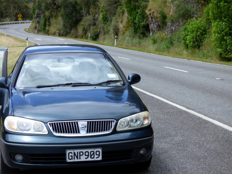 An old, blue-green Nissan bluebird standing on the side of a windy road