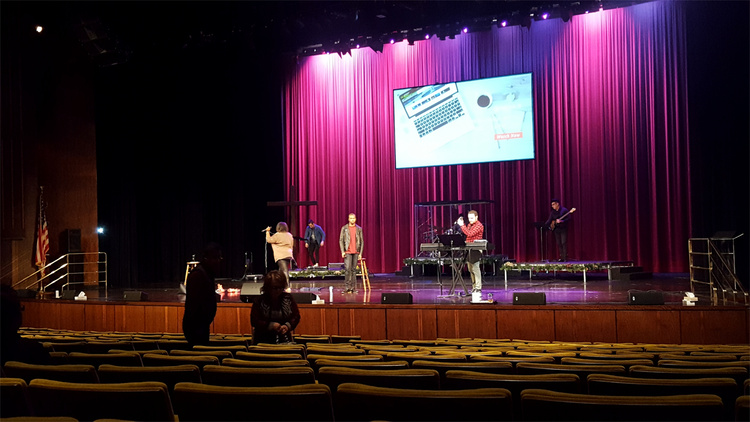A large, theatre-like auditorium with rows of empty seats and a band preparing their equipment in front of a red velvet curtain