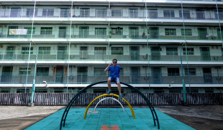 Jan sitting on an arched metal playground construction in front of tightly packed residential balconies