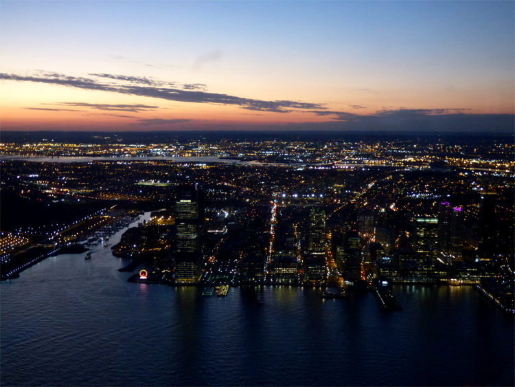 Lights of a city situated by a riverside at dusk, under an orange sunset sky