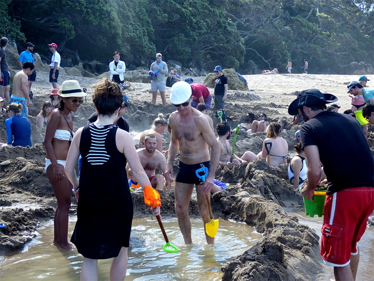 A crowd of adults in swimwear standing and sitting in pools built from sand, holding brightly coloured plastic buckets and shovels