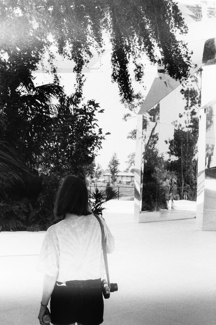 A woman with a camera around her shoulder standing in a bright space with plants and mirror-pillars