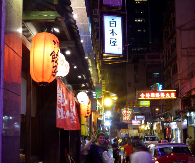 A street scene at night with various different neon signs and lanterns, with Jan making a 'peace' sign at the camera